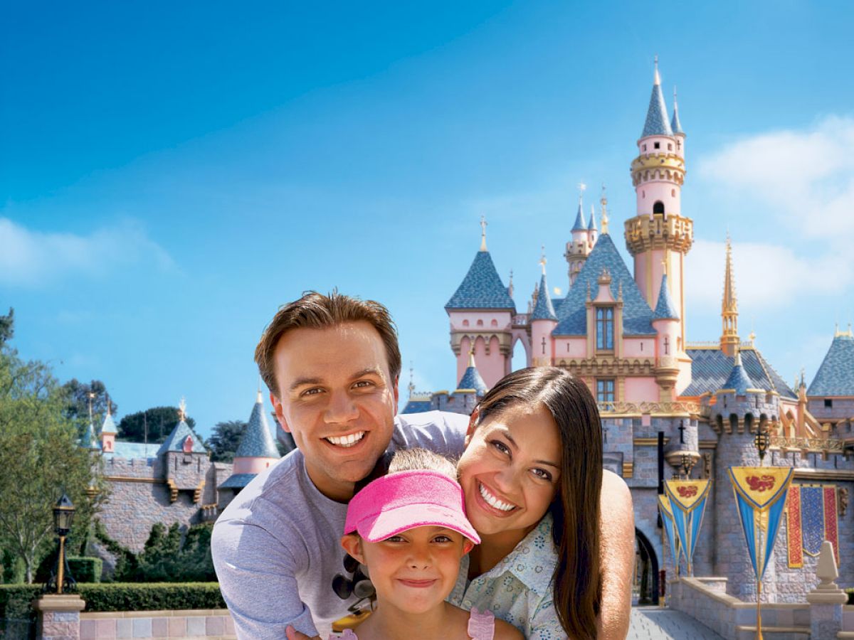 A smiling family poses in front of a castle at an amusement park, with the man and woman embracing a young girl in a pink hat, all looking happy.