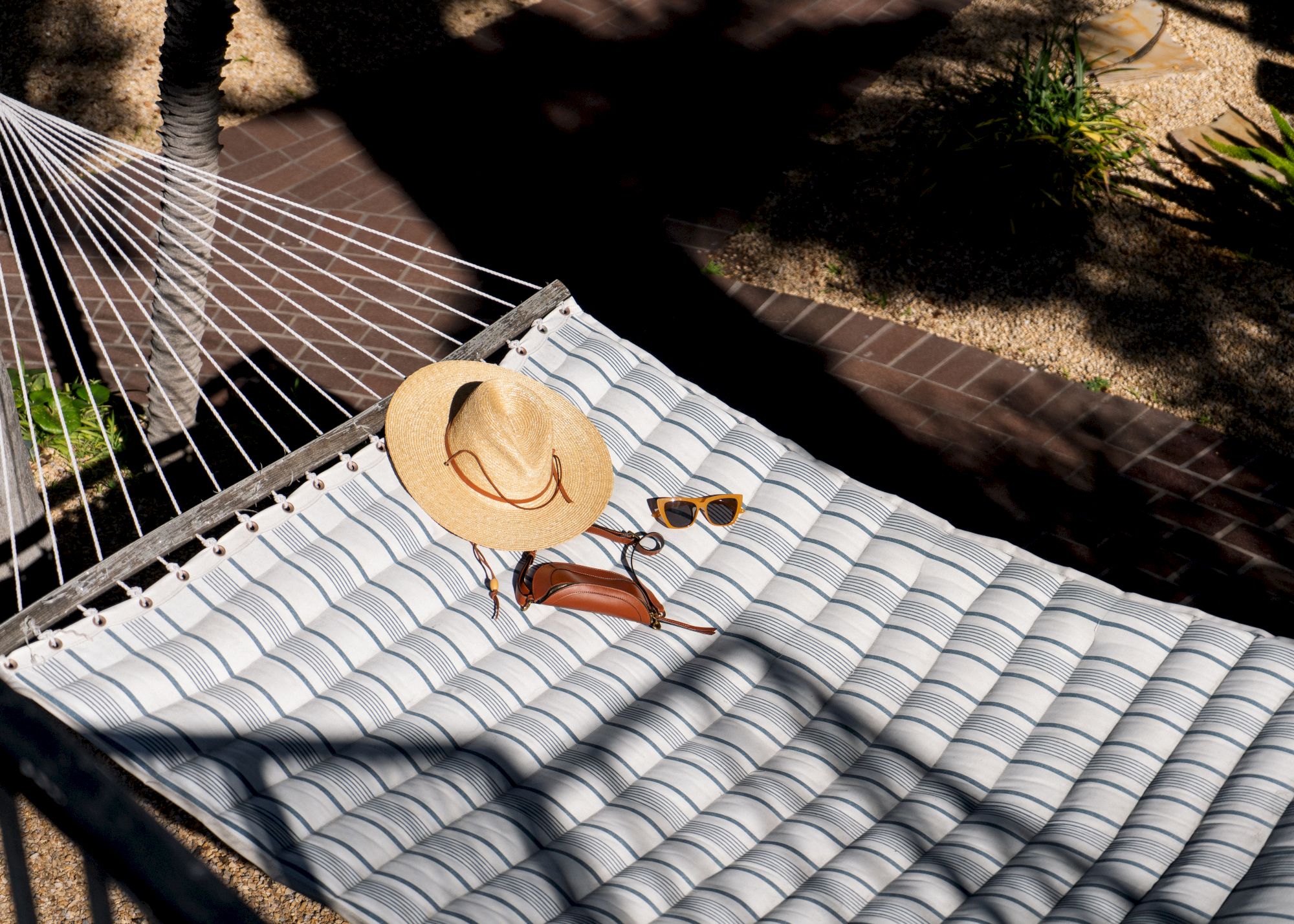 A hammock with a woven design holds a straw hat, sunglasses, and a small leather bag, basking in the dappled sunlight.