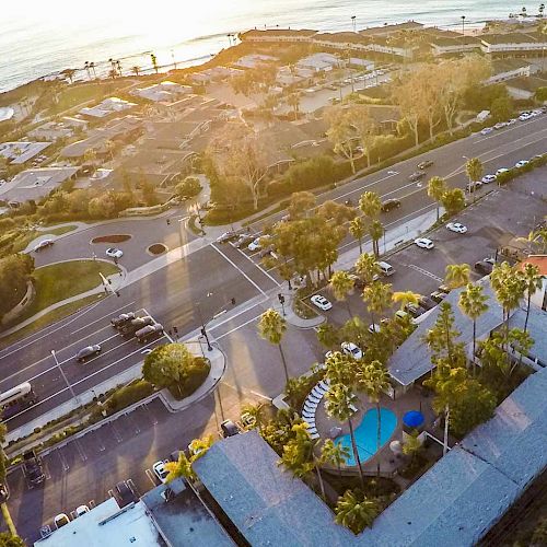 An aerial view of a coastal area with roads, buildings, a swimming pool, and greenery, leading to a beach and ocean with scattered clouds at sunset.