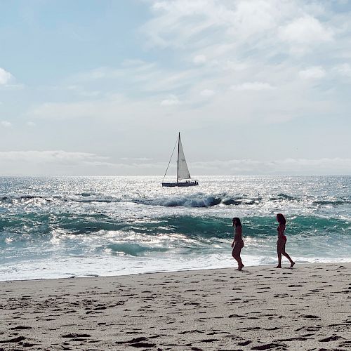 Two people walk along a sandy beach with gentle waves lapping the shore; a sailboat floats in the distance under a partly cloudy sky.