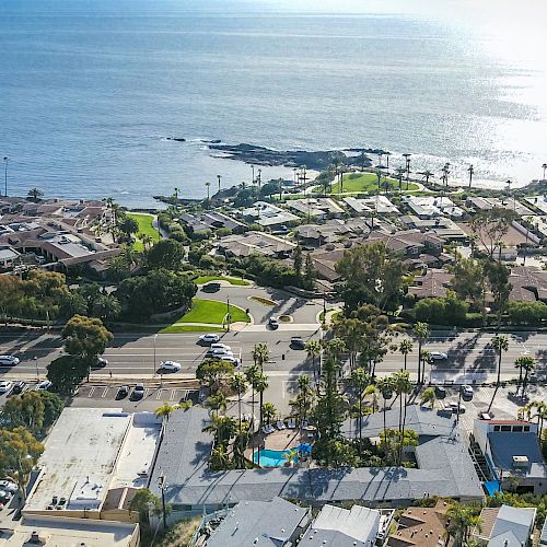 An aerial view of a coastal neighborhood with houses, roads, palm trees, and a glimpse of the ocean in the background with the shoreline visible.