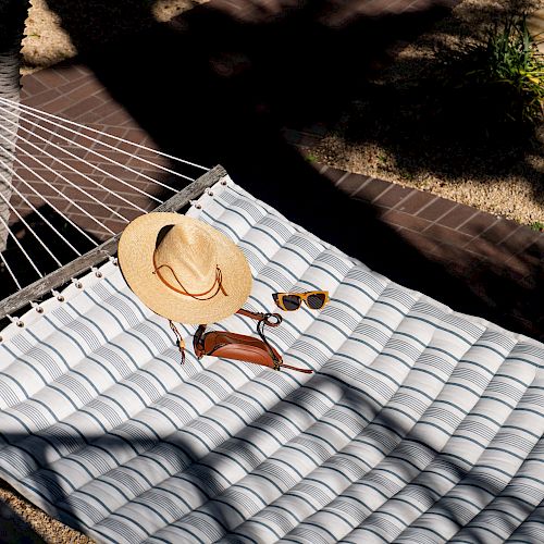 A striped hammock with a straw hat, sunglasses, and a small brown bag, casting shadows and sunlight.