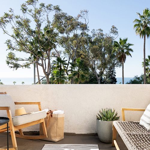 A cozy outdoor patio with chairs, a small table, potted plants, and a view of trees and the ocean in the distance ends the sentence.