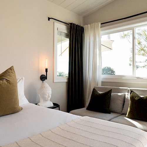 A cozy bedroom features a bed with green pillows, a couch with matching pillows, a window with a view, and a small Buddha statue.