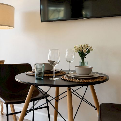 A small dining area with a round table, two chairs, place settings, and a flower vase, next to a floor lamp and beneath a wall-mounted flat-screen TV.