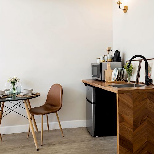 This image shows a small kitchen area with a dining table, two chairs, a sink, utensils, a microwave, and a mini-fridge on wooden countertops.