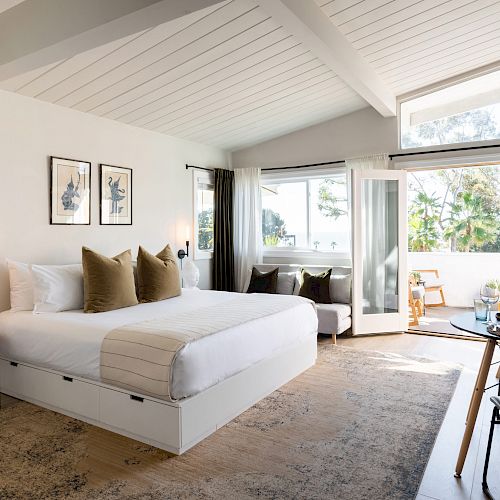 A modern bedroom with a large bed, decorative pillows, art on the walls, a rug, and a table with chairs near windows.