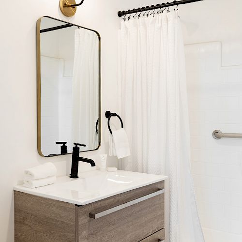 Modern bathroom with a wooden vanity, rectangular mirror, black faucet, wall sconce, white shower curtain, and towel hanging on a hook.