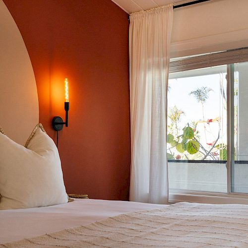 A cozy bedroom with a made bed, pillows, a wall lamp, and a large window offering a view of palm trees and plants outside.