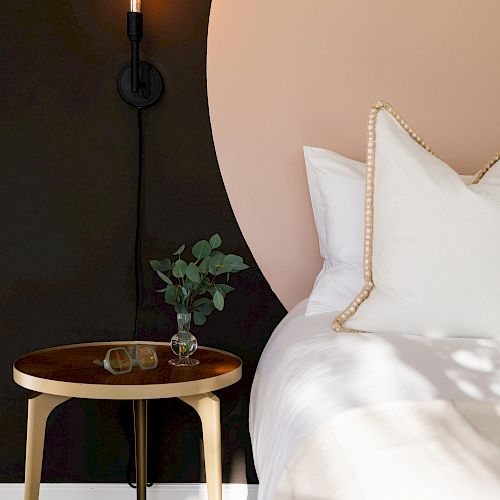 A modern bedroom setup with a round side table, minimalistic wall light, and part of a bed with white pillows and sheets, featuring a two-tone wall.