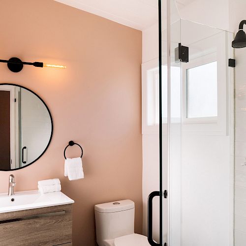 A modern bathroom features a vanity with a round mirror, towel holder, toilet, and glass shower enclosure with black fixtures and white walls.