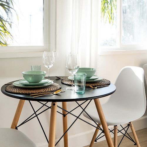 A small dining table set for two with green dishware, wine glasses, and placemats. White chairs, large windows, and bright sunlight complete the setting.