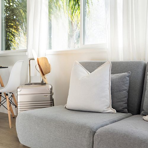 The image shows a cozy living room with a gray sofa, a white pillow, a small wooden table, a chair, and large windows with white curtains.