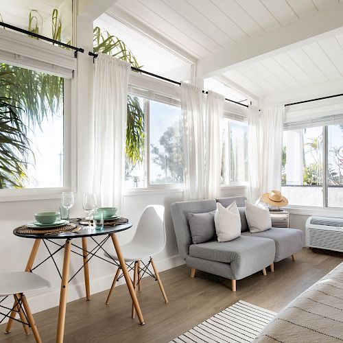 This image depicts a bright, cozy living area with large windows, white curtains, a small dining table, modern chairs, a gray sofa, and wooden flooring.