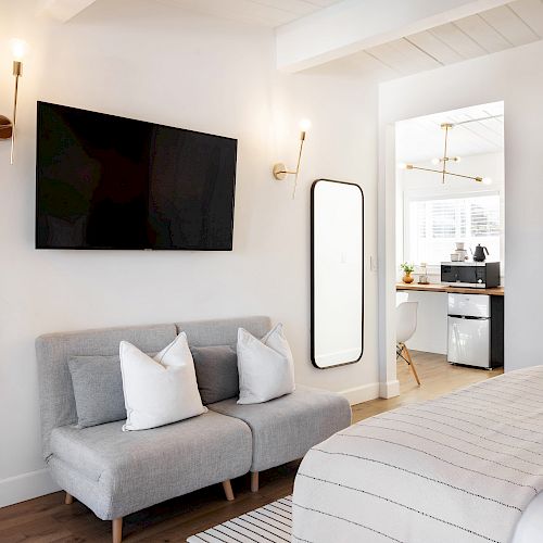 A modern bedroom with a wall-mounted TV, small sofa, floor mirror, and view into a bathroom with a mini-fridge and white decor ending the sentence.