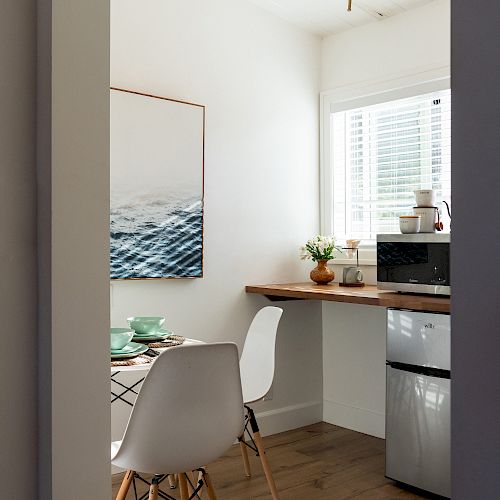 The image depicts a small kitchen or dining area with white chairs, a wooden table, a small fridge, a microwave, a window, and a painting.