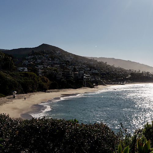 A coastal view features a sandy beach, ocean waves, and hills with houses, under a clear sky with the sun shining.