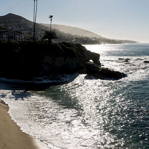 A scenic coastal view with a sandy beach, rocky cliffs, palm trees on a hill, and sunlight reflecting off the ocean waves.