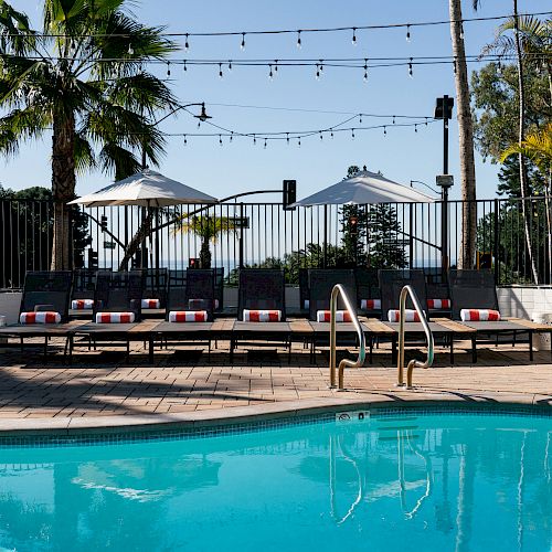 The image shows a swimming pool with sun loungers, umbrellas, string lights, and palm trees, suggesting a relaxing outdoor area.