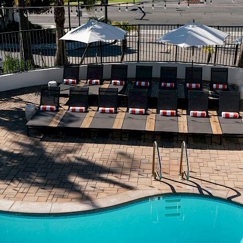 The image shows a swimming pool with lounge chairs and umbrellas beside it, located near a road with palm trees and traffic lights in the background.