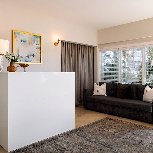 A cozy living room featuring a dark gray sofa, a large rug, a white cabinet, wall art, plants, and ample natural light from the windows.