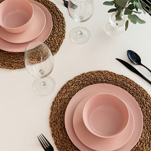 A table set for two with pink dishes, glasses, black cutlery, and woven placemats; a small green plant is in the background.