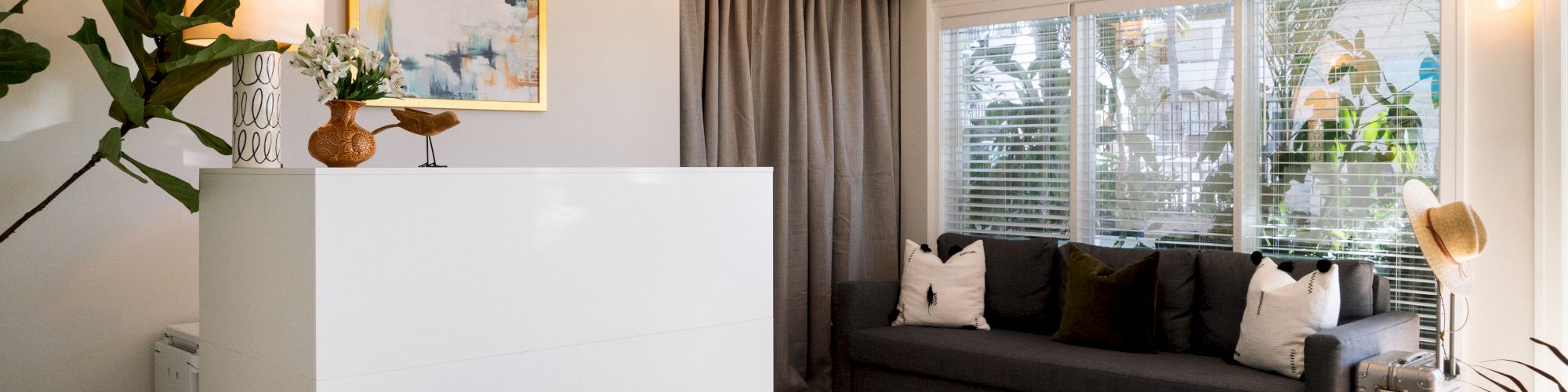 A cozy living room with a gray sofa, abstract wall art, houseplants, a white partition, and a large rug, illuminated by natural light from the windows.