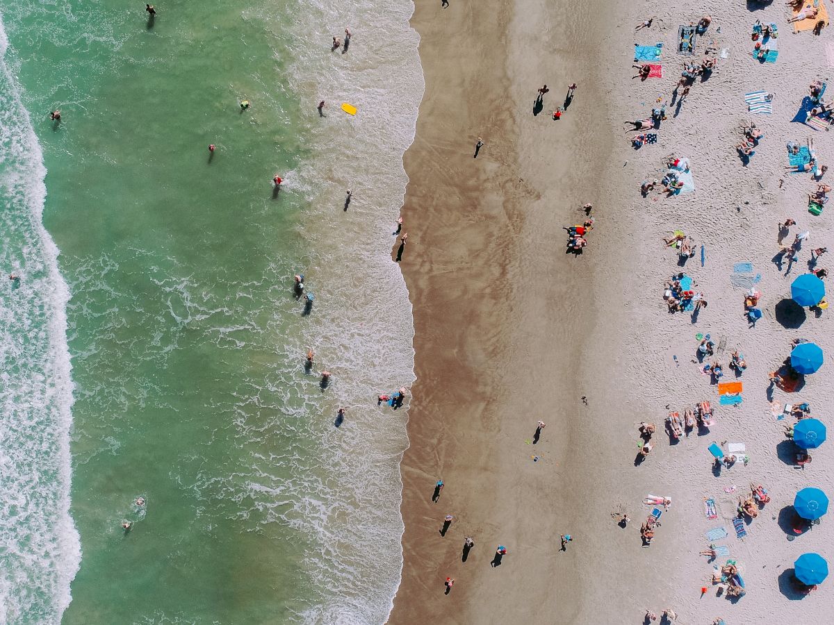 An aerial view of a crowded beach shows people on the sand and in the water, with umbrellas and beach items scattered around.