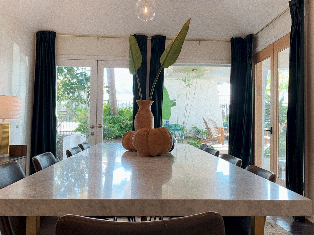 A modern dining room with a long table, chairs, a plant centerpiece, pendant lights above, and windows with dark curtains letting in natural light.