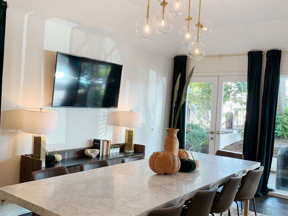 A modern dining room with a large marble table, black chairs, a chandelier, a mounted TV, table lamps, and decorative vases. Large windows with curtains.