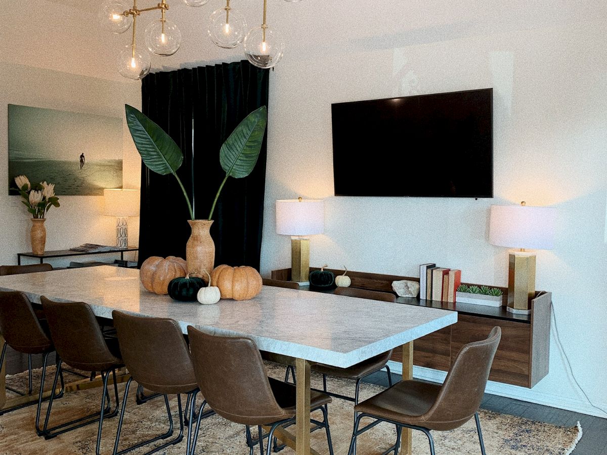 A modern dining room with a long table, brown chairs, a large plant centerpiece, wall art, a rug, a TV, and a chandelier with hanging bulbs.