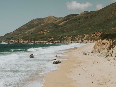 A sandy beach with waves crashing on the shore, rocky cliffs, and mountains in the background under a partly cloudy sky.