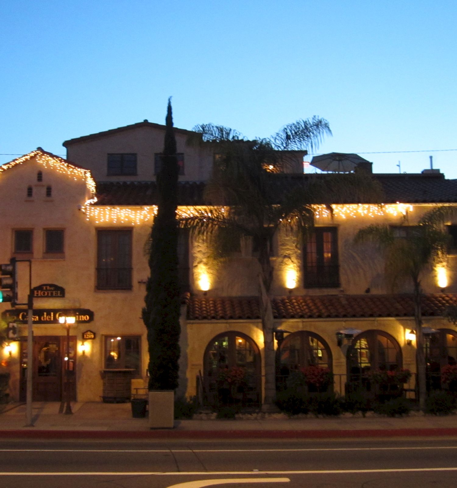 The image shows a building with an illuminated facade and decorative lights, featuring arches, palm trees, and a clear evening sky.