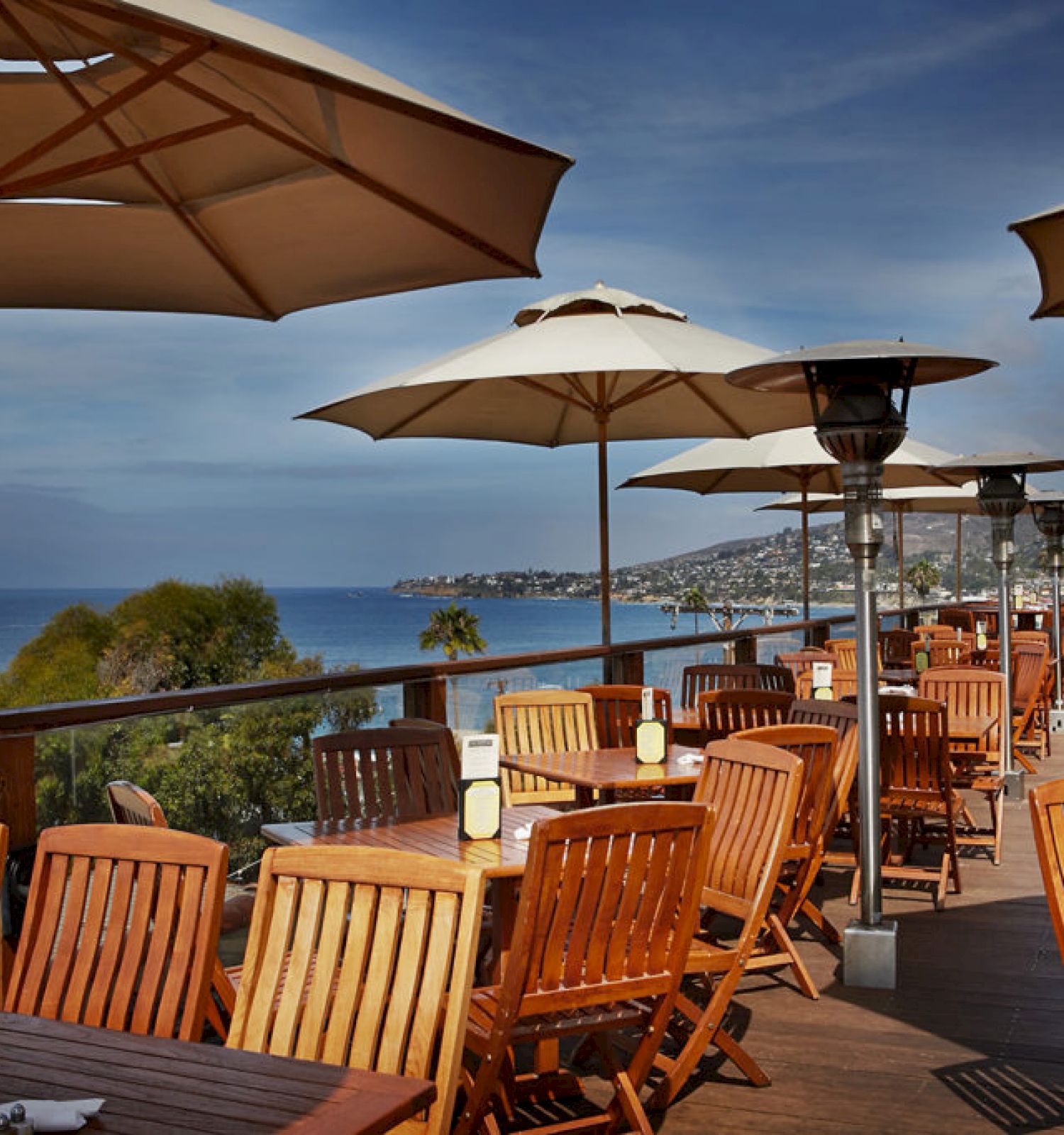 This image shows an outdoor restaurant patio with wooden tables and chairs, shaded by umbrellas. The patio overlooks a scenic ocean view.