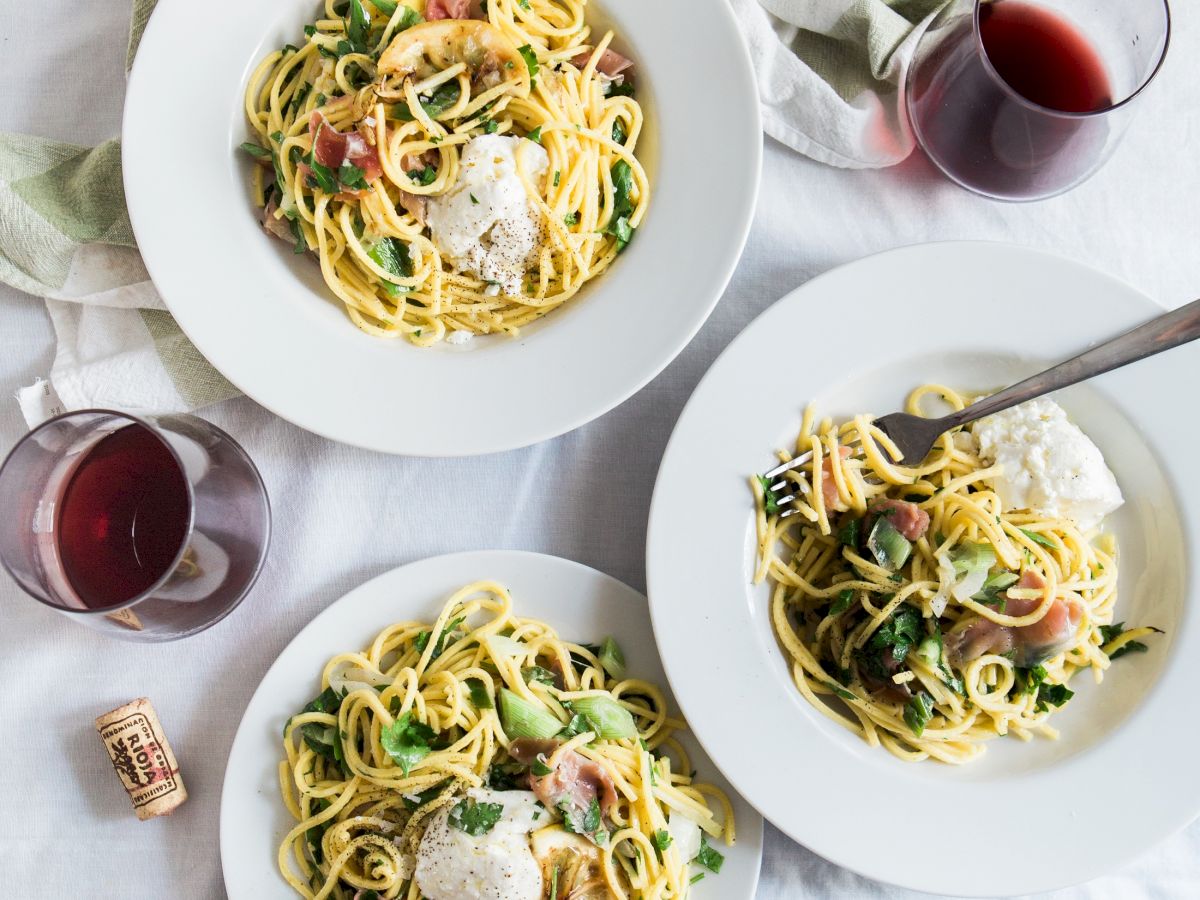 Three plates of pasta with greens and cheese, accompanied by two glasses of red wine and a cork, all on a white tablecloth.