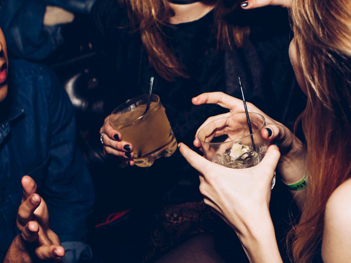 Three people are in conversation holding drinks, with one person touching another's face and everyone smiling, suggesting a lively social scene.