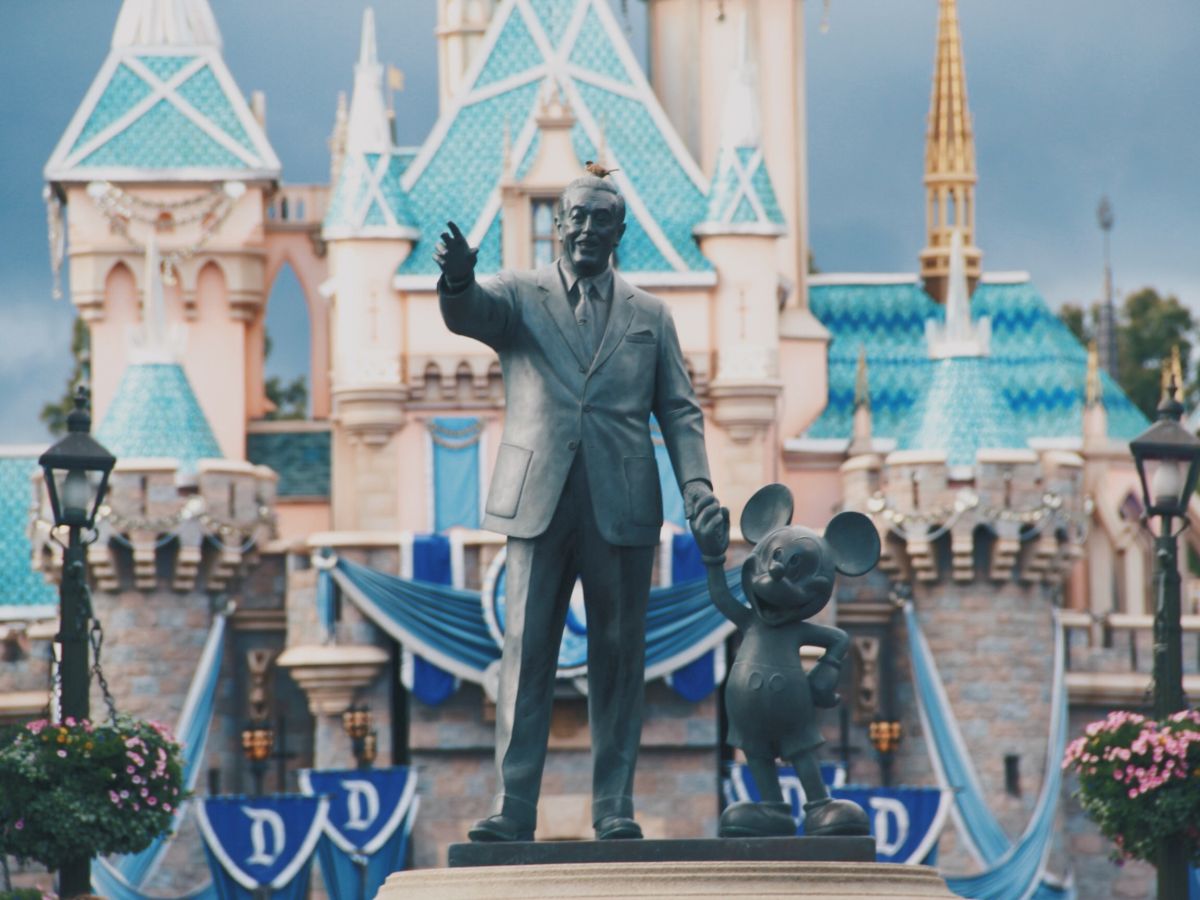 A statue of a man and a mouse character in front of a castle with blue turrets and decorative banners in an amusement park.