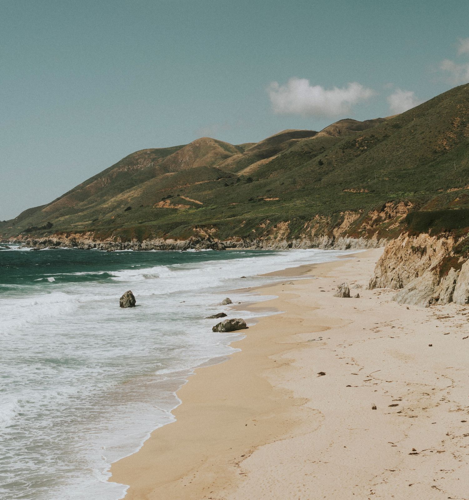 A serene beach scene with sandy shores, gentle waves, and rocky cliffs against a backdrop of green hills under a partly cloudy sky.