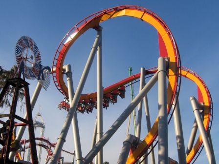 The image shows a vibrant orange and yellow roller coaster with loops and twists set against a clear blue sky.