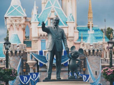 A statue of a man and a mouse character stands in front of a fairy-tale castle, surrounded by flowers and banners displaying the letter 